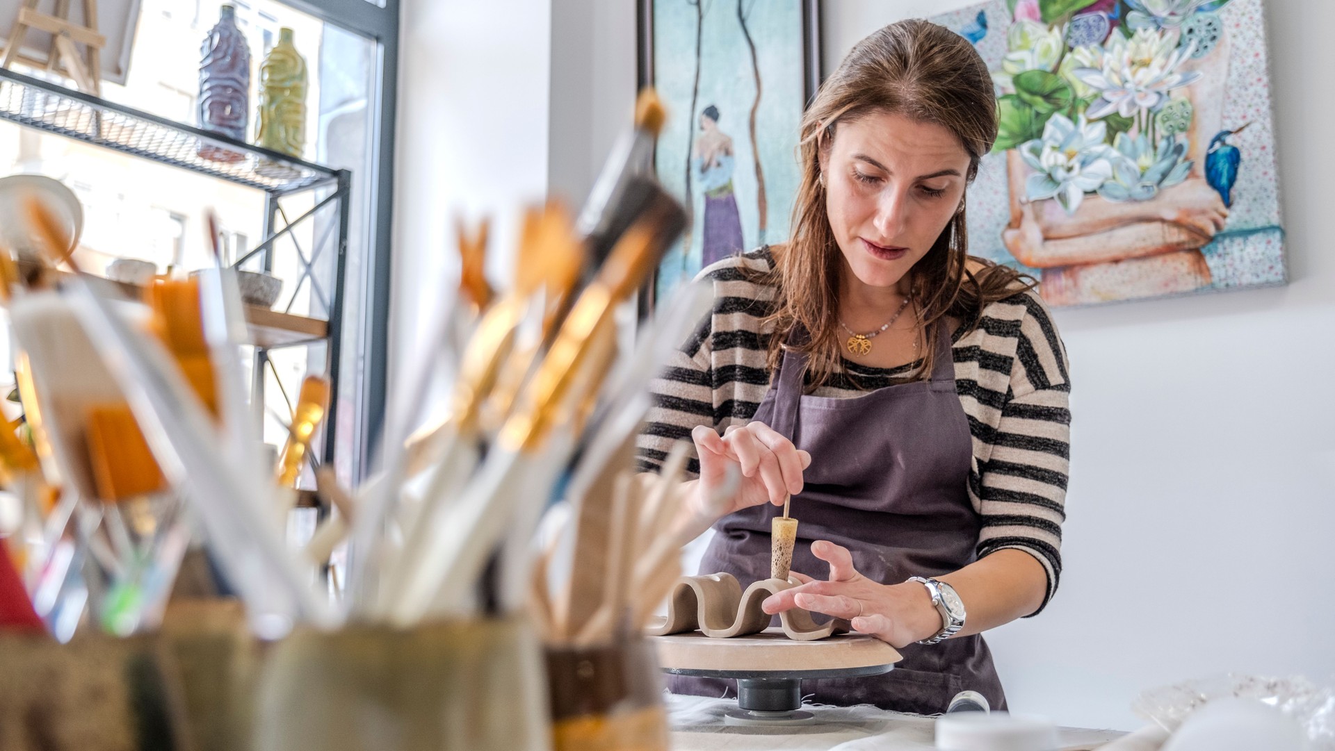A shot of a middle-aged woman artist shaping clay with sculpting tools at a table in an art studio, with an environment adorned with artistic paintings in the background