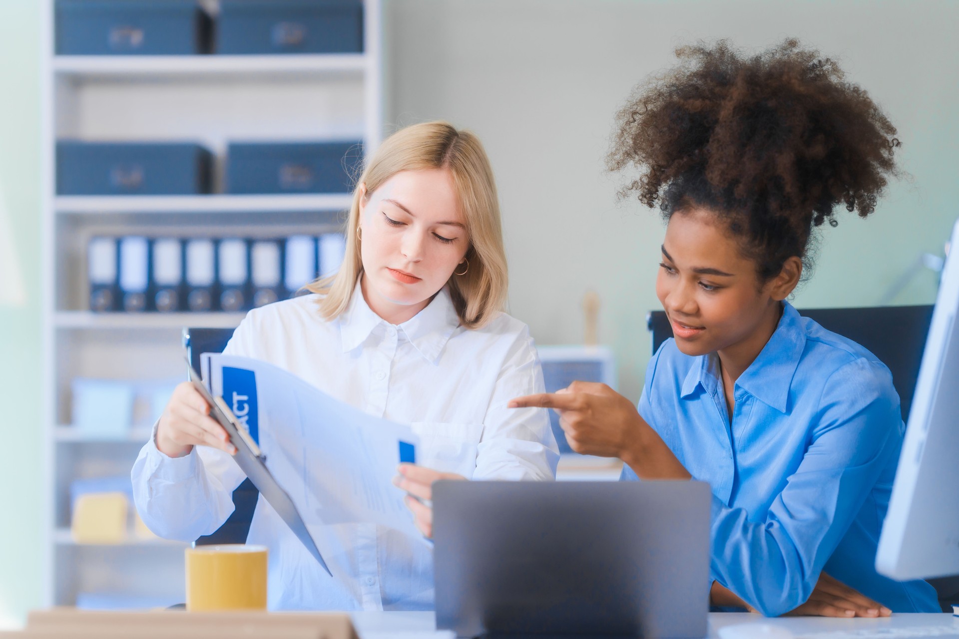 Young African American woman and Russian woman work together in business office, consulting on financial charts using desktop PC and tablet. They discuss strategies and collaborate as colleagues.