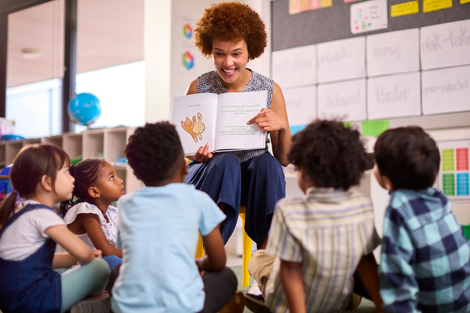 Female Teacher Reads To Multi-Cultural Elementary School Pupils Sitting On Floor In Class At School